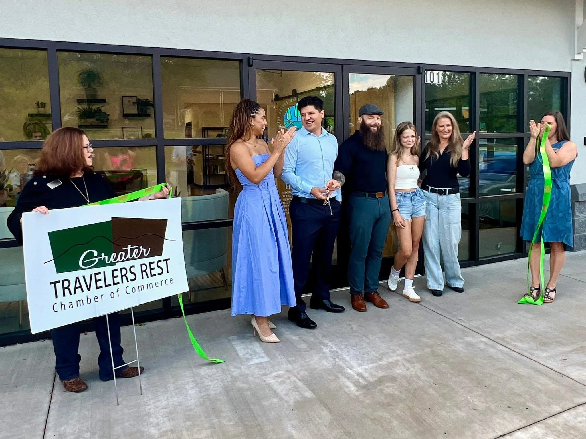 Group of people standing outside in front of a building during a ribbon-cutting event. Two women are holding a large sign that reads 'Greater Travelers Rest Chamber of Commerce,' with one woman on each end of the ribbon. The group is clapping and smiling, celebrating the occasion.