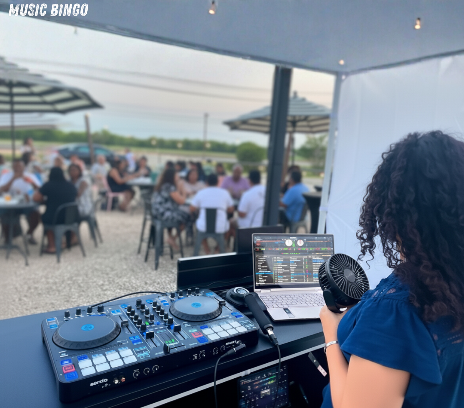 A DJ woman with curly hair wearing a blue dress operating DJ equipment with a laptop and handheld fan at an outdoor event with people sitting at tables under umbrellas.