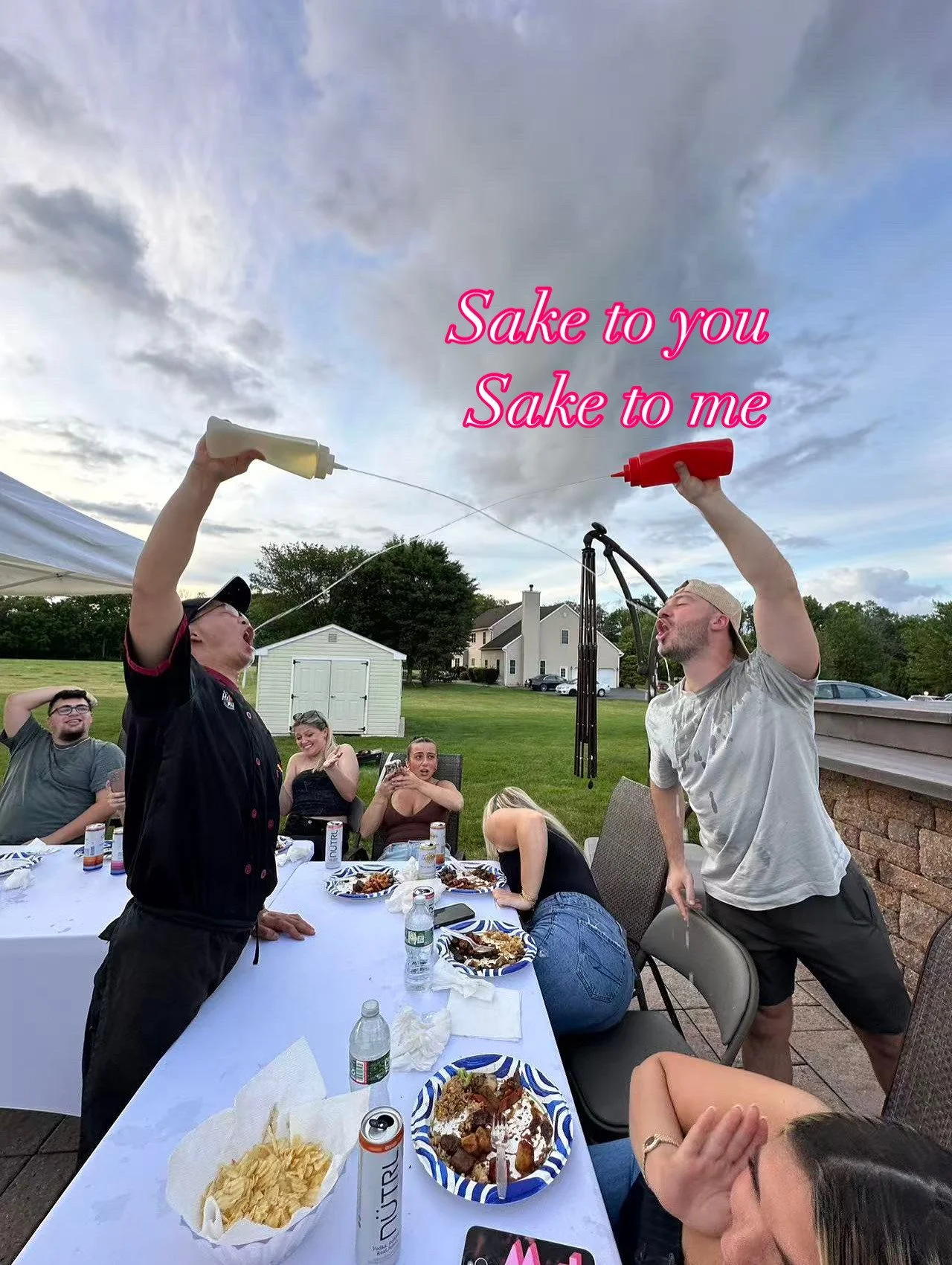 People at an outdoor gathering sharing a humorous moment with two men pretending to spray drinks into each other's mouths, with the caption 'Sake to you Sake to me' in pink text.