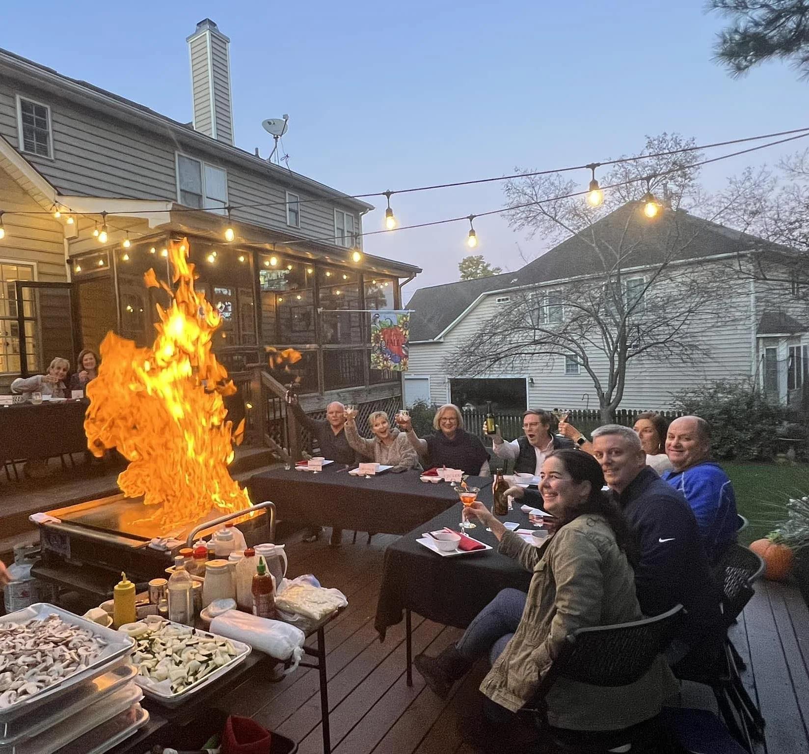 A group of people sitting at a dining table outdoors, celebrating with drinks, while a large flame from a grill or fire pit illuminates the scene, with string lights overhead and a residential neighborhood in the background.