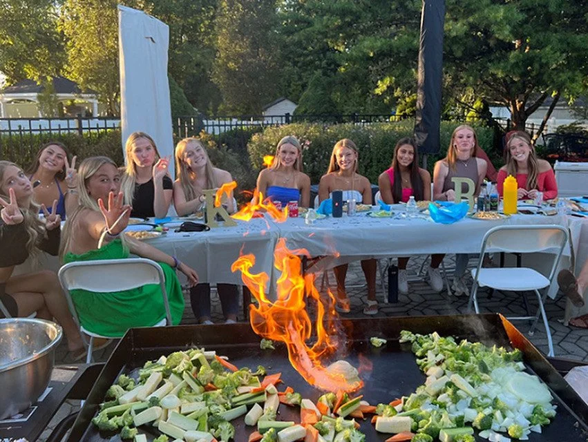 Group of young women sitting at a long table outdoors during a cookout or party, with a flaming grill and vegetables in the foreground.