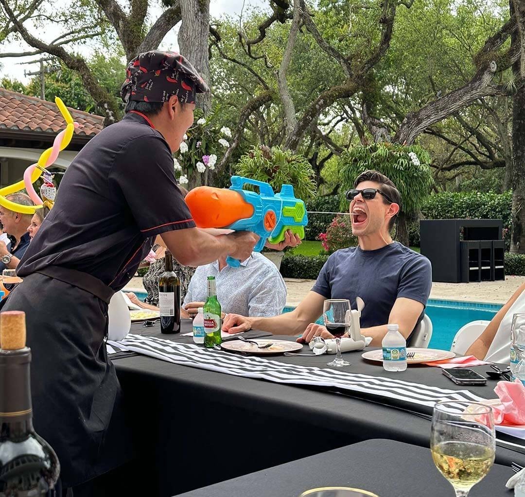 A man in sunglasses sitting at a table with food and drinks, with his mouth open as if laughing or shouting, as a woman in a black chef's outfit with a bandana squirts him with a water gun during an outdoor gathering near a swimming pool.