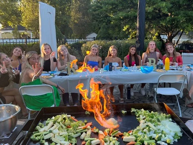 A group of young girls and women sitting at a long outdoor table celebrating with a barbecue grill in front of them, with vegetables cooking on the grill and flames visible.