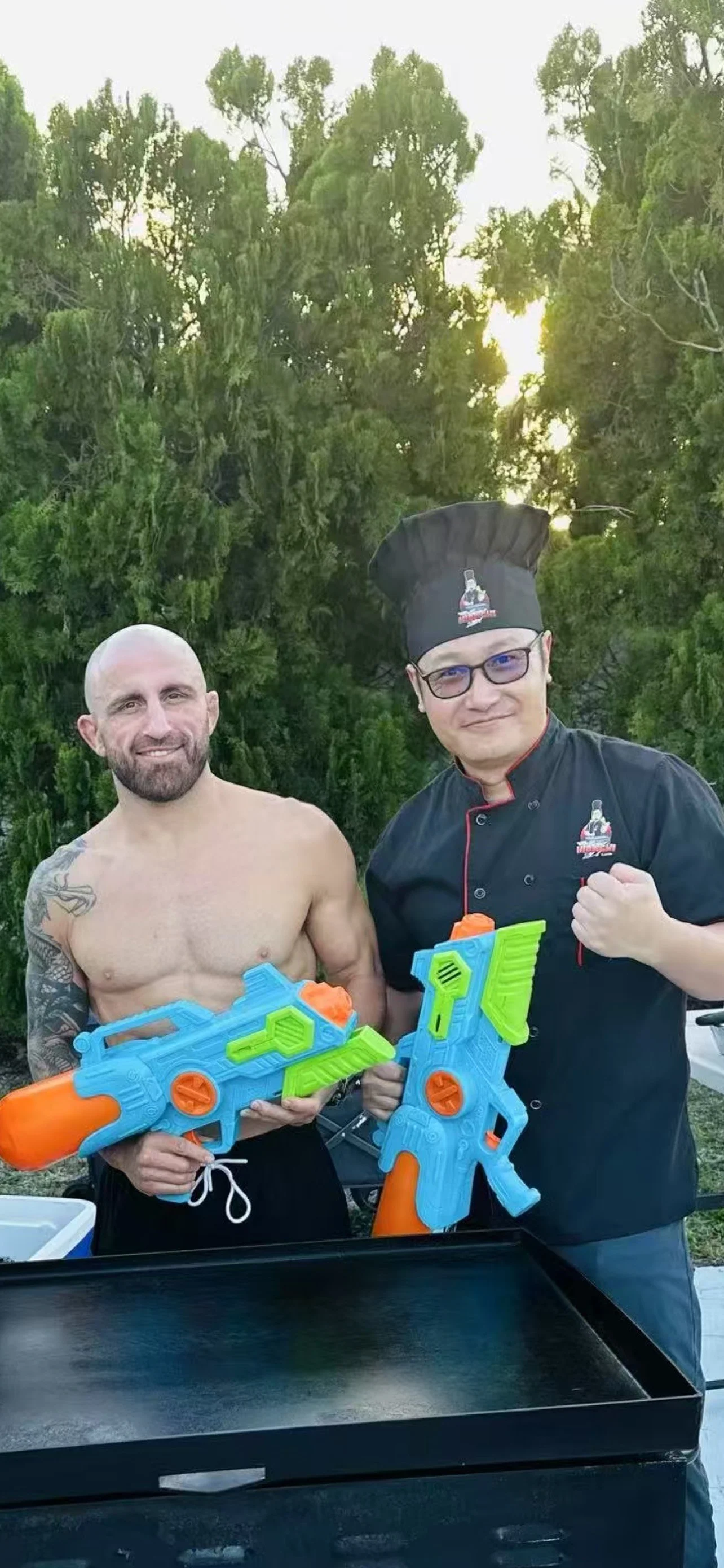 Two men holding toy water guns outdoors, one shirtless and the other in a chef's uniform and hat, with green trees in the background.