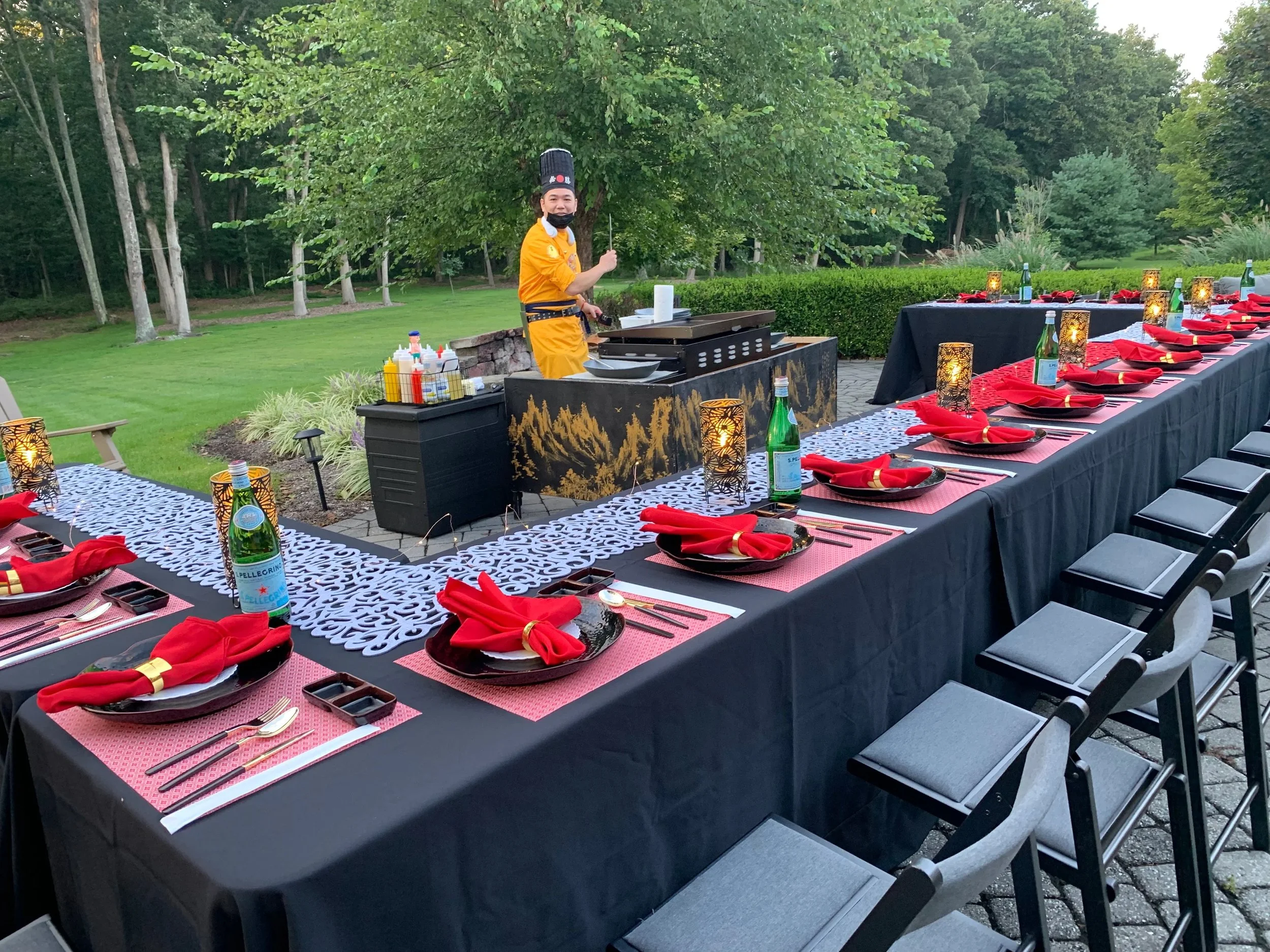 An outdoor dining setup with a long table covered in black and red table settings, red napkins, lit candles, and glass bottles of sparkling water, with a chef in yellow attire preparing food at a black and gold-painted grill, surrounded by green tree