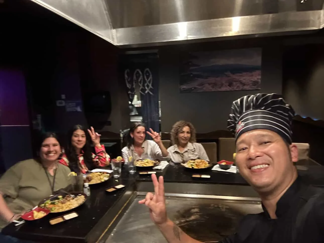 A group of four women and a chef sitting at a table in a restaurant with plates of food, two of them making peace signs, and the chef taking a selfie.