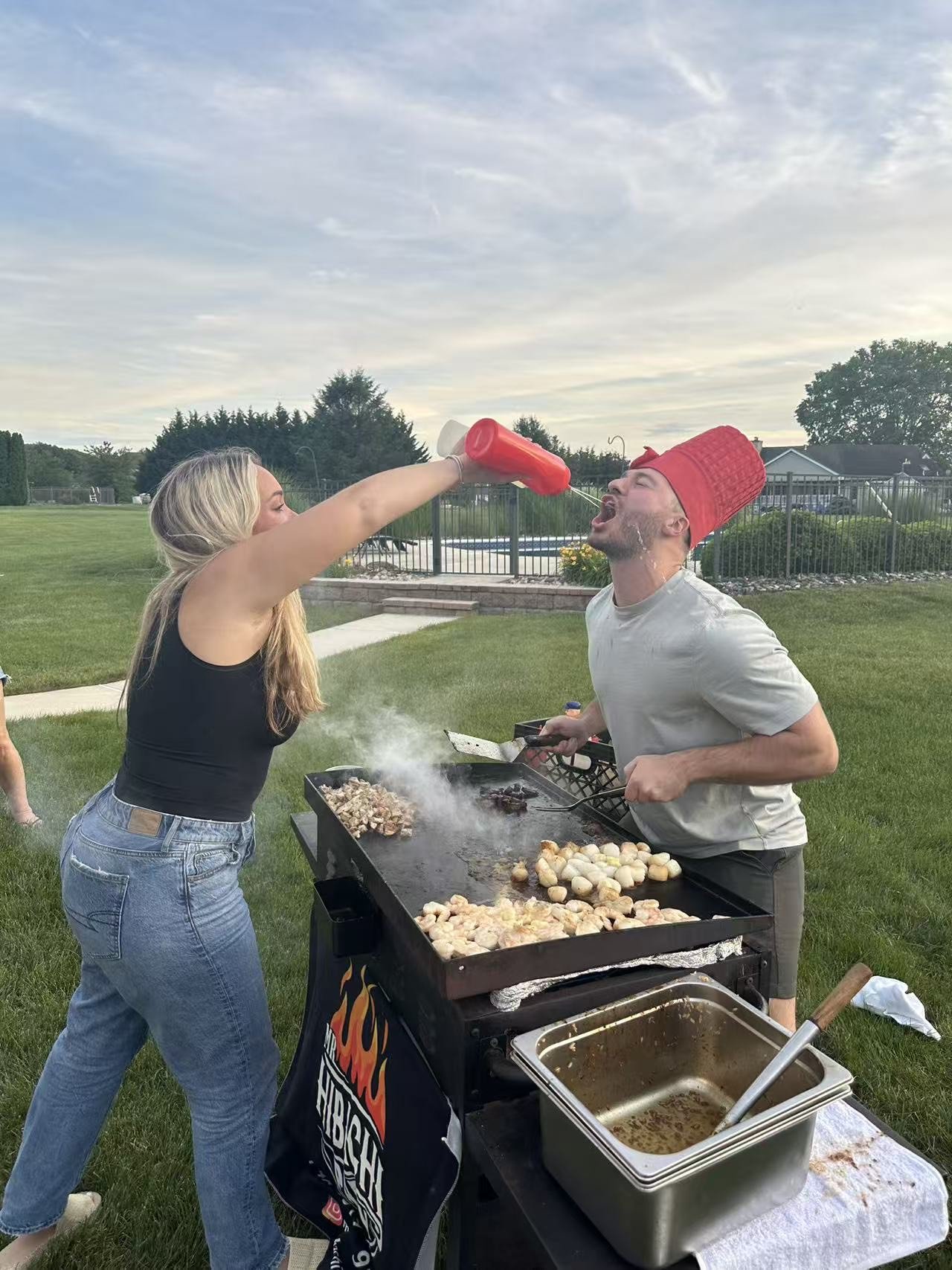A woman spray-feeds a man with ketchup during a barbecue outdoors.