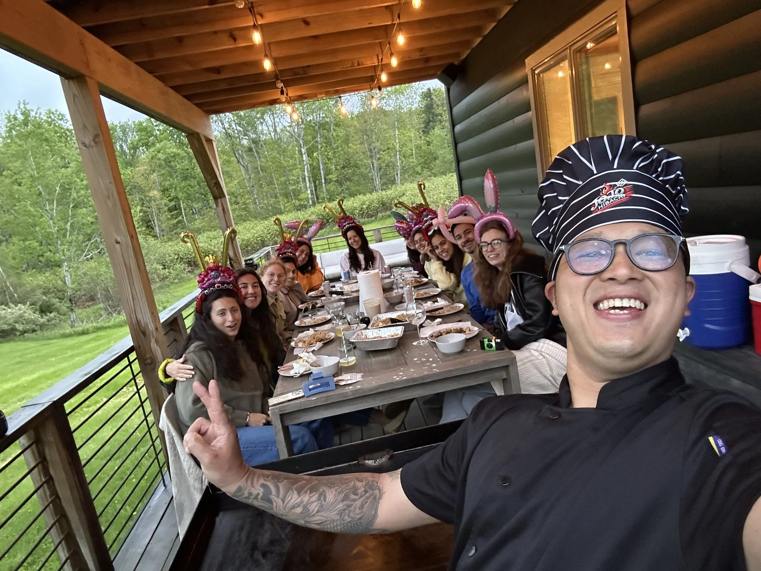 Group of people sitting at a long dining table on a covered porch, wearing pink bunny ear headbands, celebrating Easter, with trees and greenery in the background.