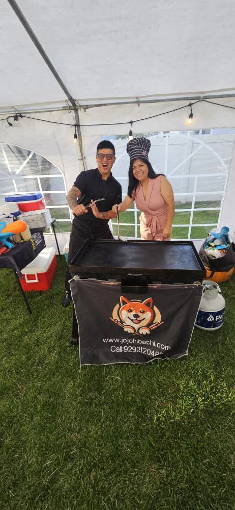 Two people, a man and a woman, are smiling and posing with a grill inside a white tent at an outdoor event. The man is holding tongs, and the woman is wearing a chef's hat and pink outfit. There are food and coolers nearby, and a banner with a dog lo