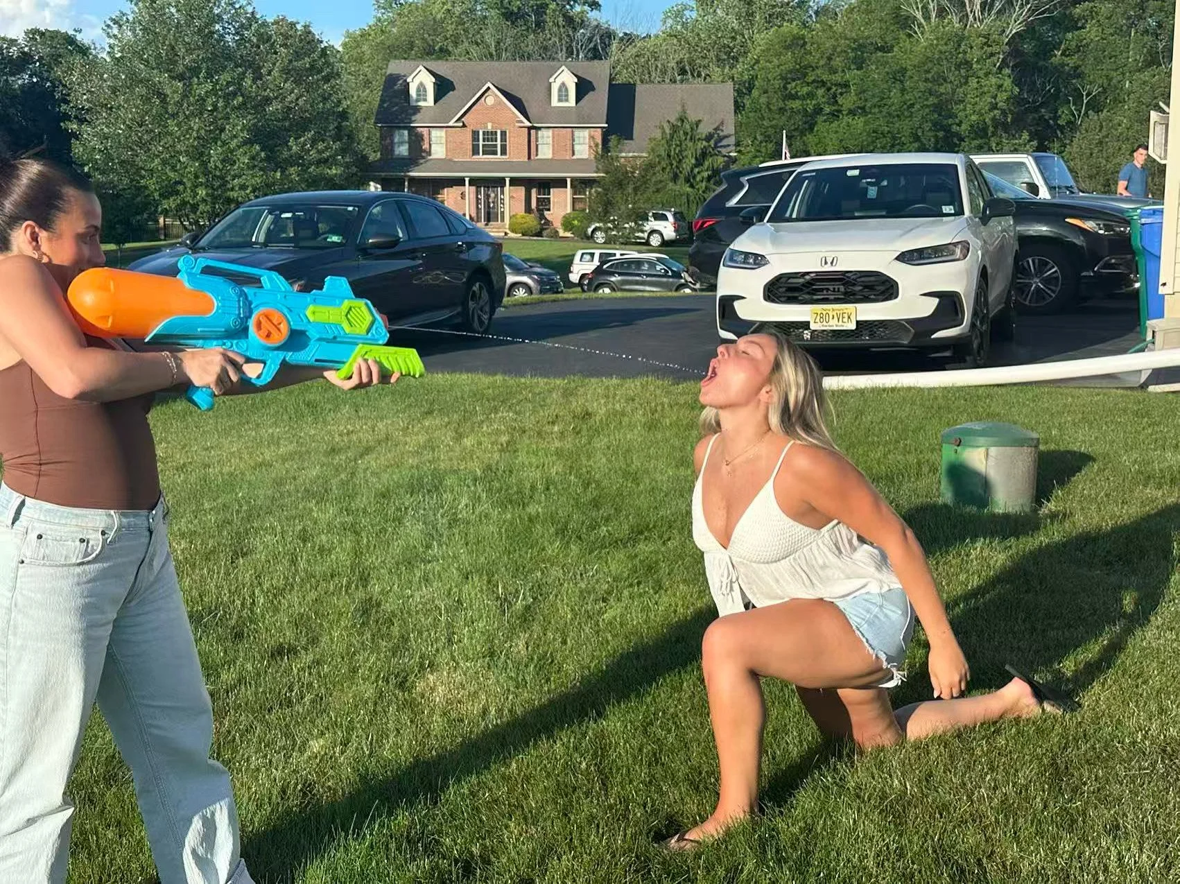 A woman kneeling on grass with her mouth open, pretending to be shot by another woman holding a large water gun, in a parking lot with cars and a house in the background.