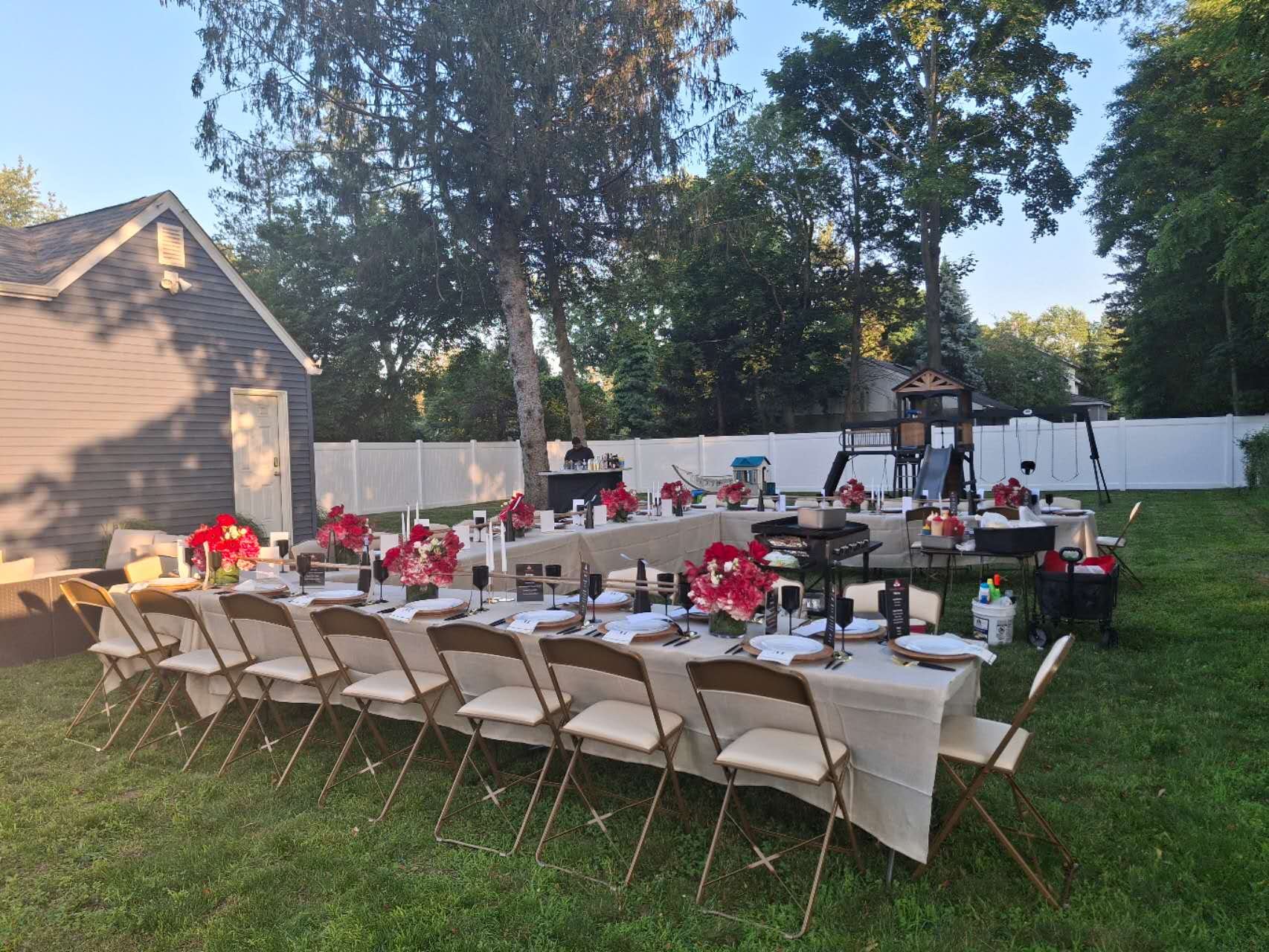Outdoor backyard party setup with a long table decorated with pink floral centerpieces and place settings, a playground in the background, and a white fence surrounding the area on a sunny day.