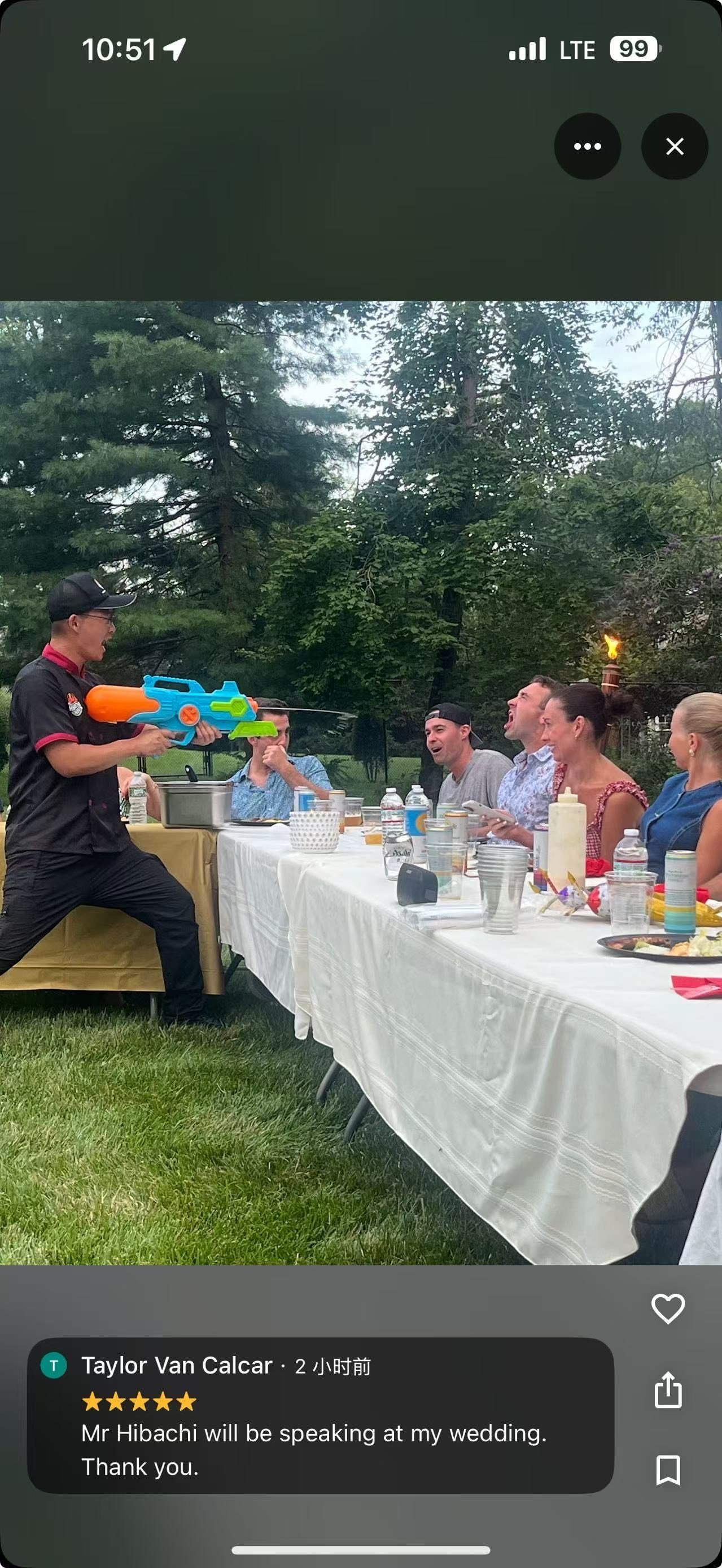A man at a wedding reception is spraying guests with a water gun. The guests, seated at a long table outdoors, are smiling and laughing, enjoying the playful moment. The table is covered with a white tablecloth and has various drinks and plates on it