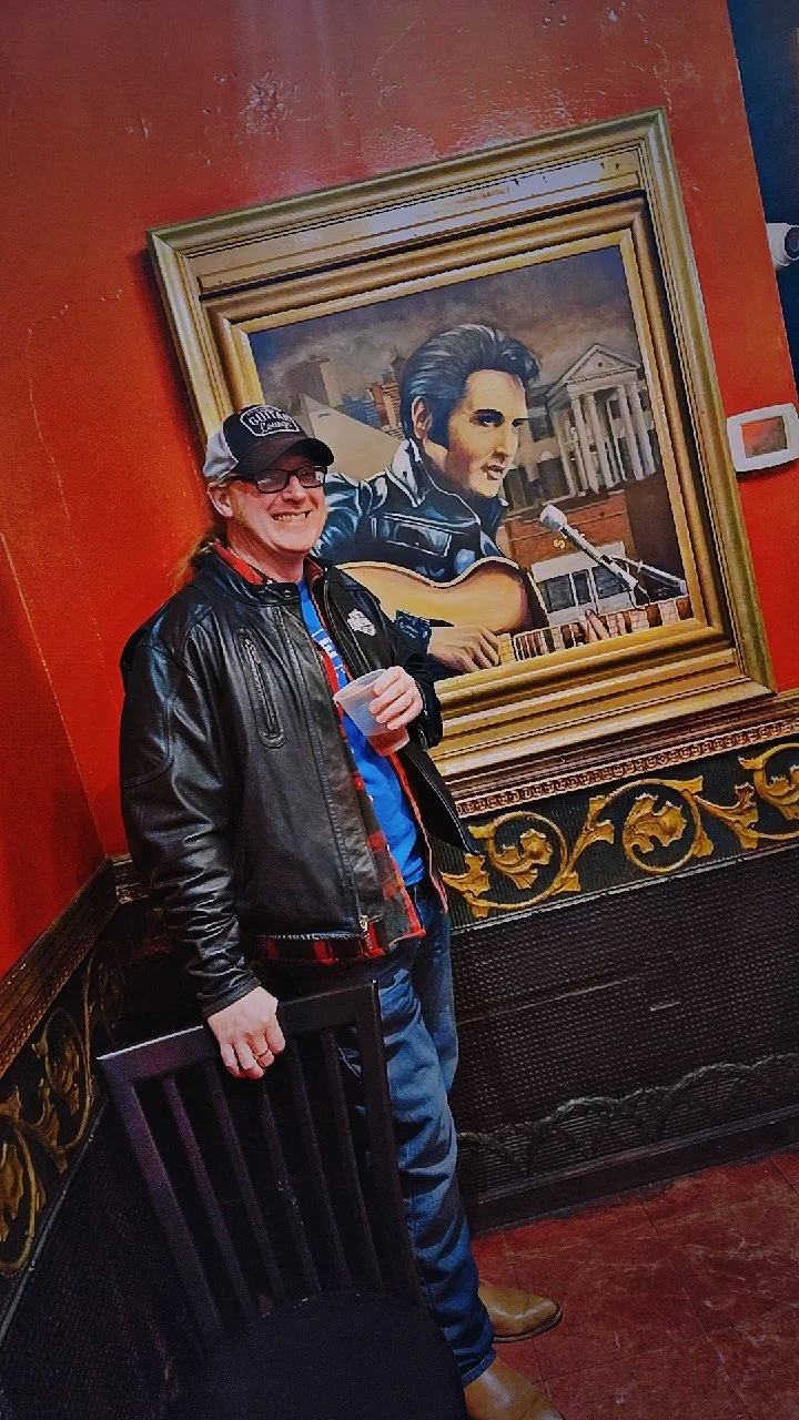 A man in a leather jacket and cap smiling, standing next to a portrait of Elvis Presley playing guitar in a room with red walls and ornate black and gold trim.