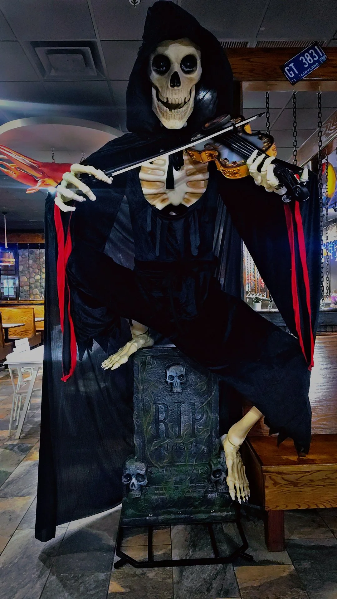 A Halloween display of a skeleton dressed as a pirate, holding a violin and a flaming sword, standing on a tombstone with skulls in a restaurant or bar.