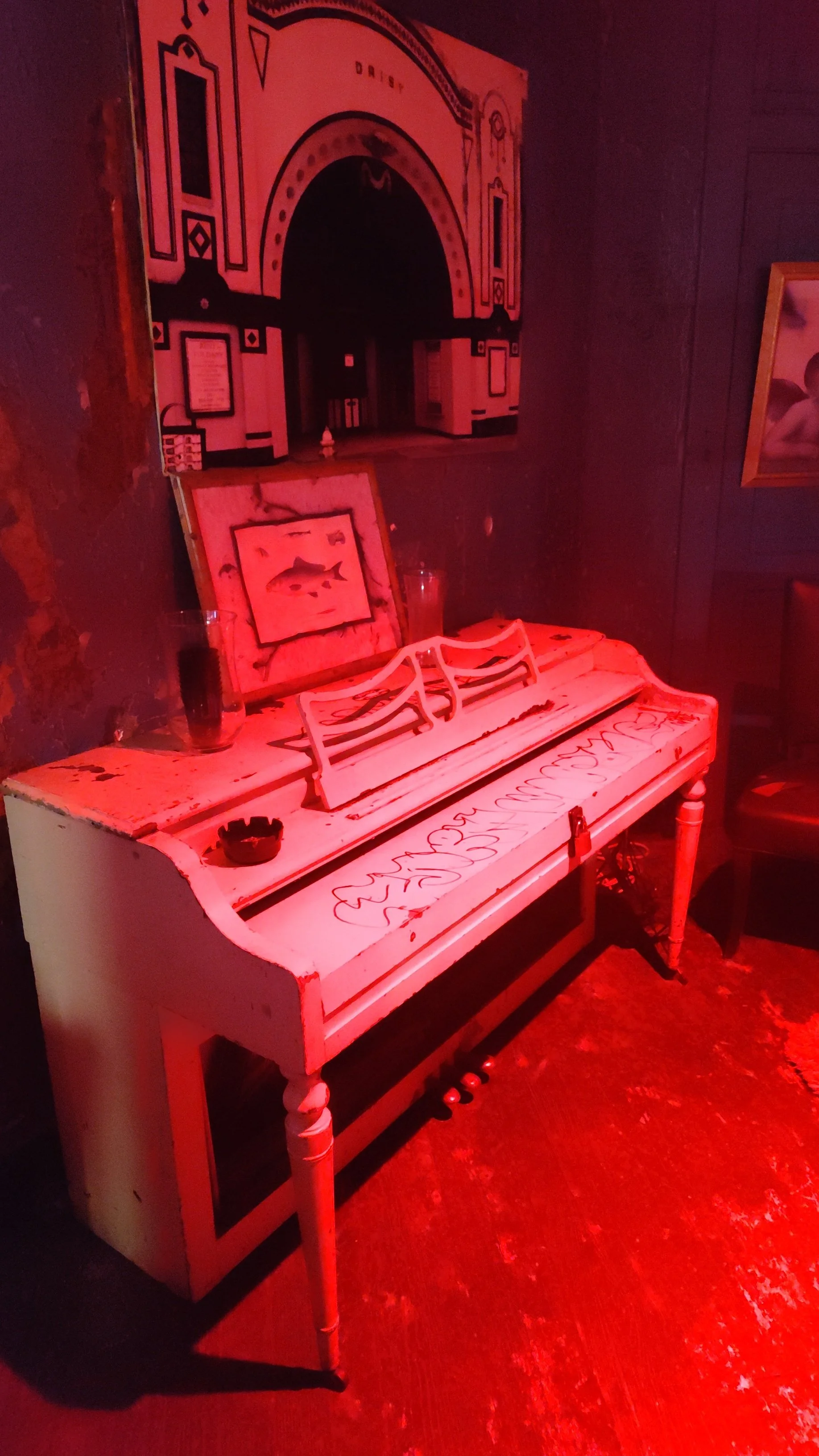 An old white piano with a music stand, decorated with red graffiti, is placed in a dimly lit room with red lighting. Behind the piano, there are framed pictures and paintings on the wall, including one of a fish and another of an abstract face.