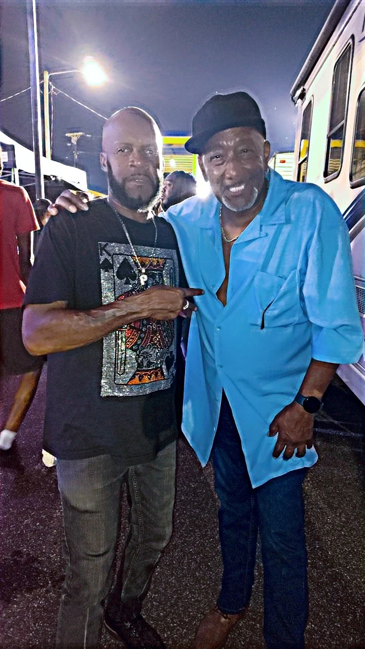 Two men standing side by side outdoors at night, one pointing at the other, with a vehicle and a bright streetlamp in the background.