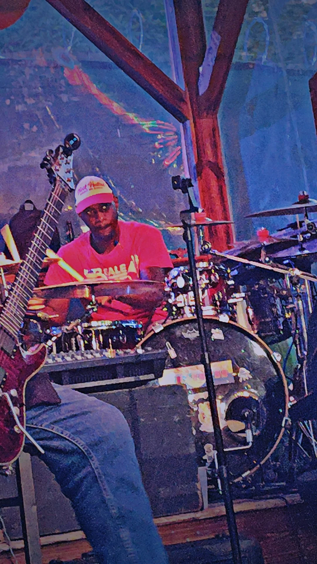 A man wearing a pink shirt and white cap sitting behind a drum set, with a guitar and microphone nearby, in a colorful, stage-like setting.