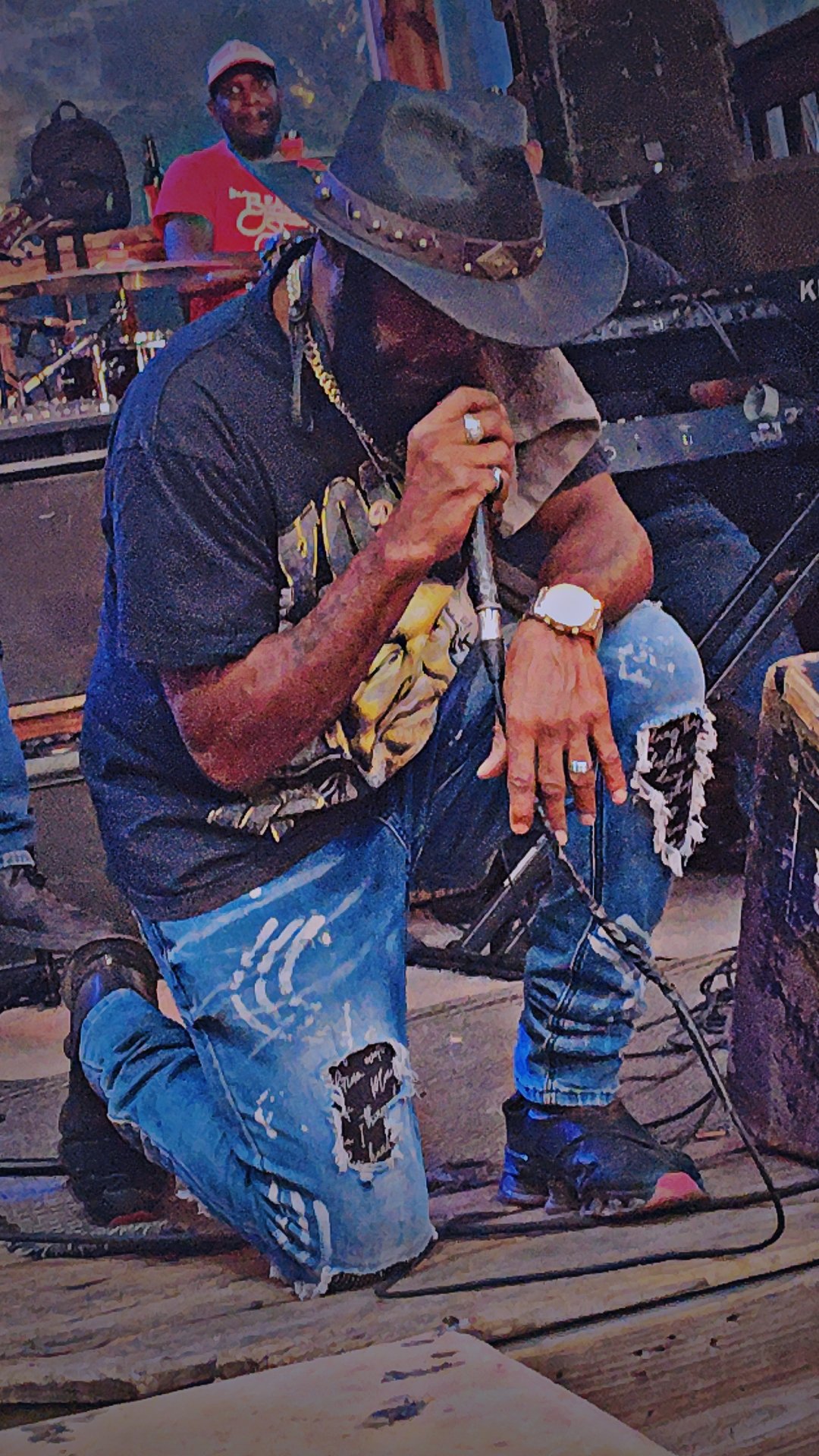 A person is kneeling on the floor, wearing a cowboy hat, sunglasses, and distressed jeans, holding a microphone and looking down. Another person in a red shirt and glasses is in the background, smiling and standing near drums.