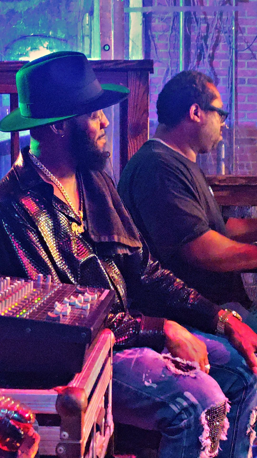 Two men sitting indoors at a dimly lit venue, one wearing a black hat, patterned jacket, and gold chain, and the other wearing glasses and a black shirt, with a music mixer in front of them.
