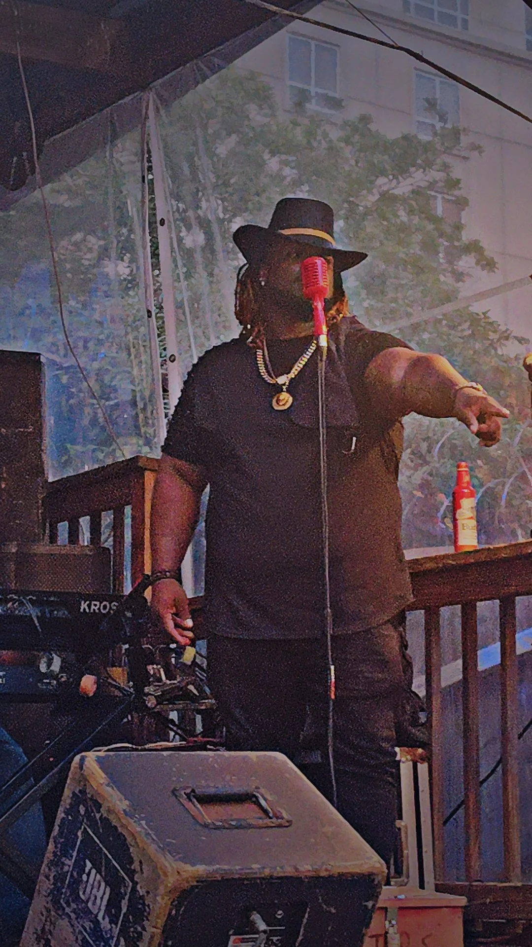 A person wearing a black hat, black shirt, and gold jewelry standing behind a microphone. The person is pointing towards the right. There is a drum nearby and a spray bottle on the wooden railing behind him. The background shows a partially visible b
