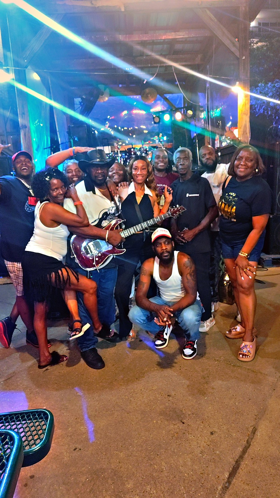 Group of people posing on a stage at a lively music event, some holding musical instruments, under colorful stage lighting in an indoor venue.