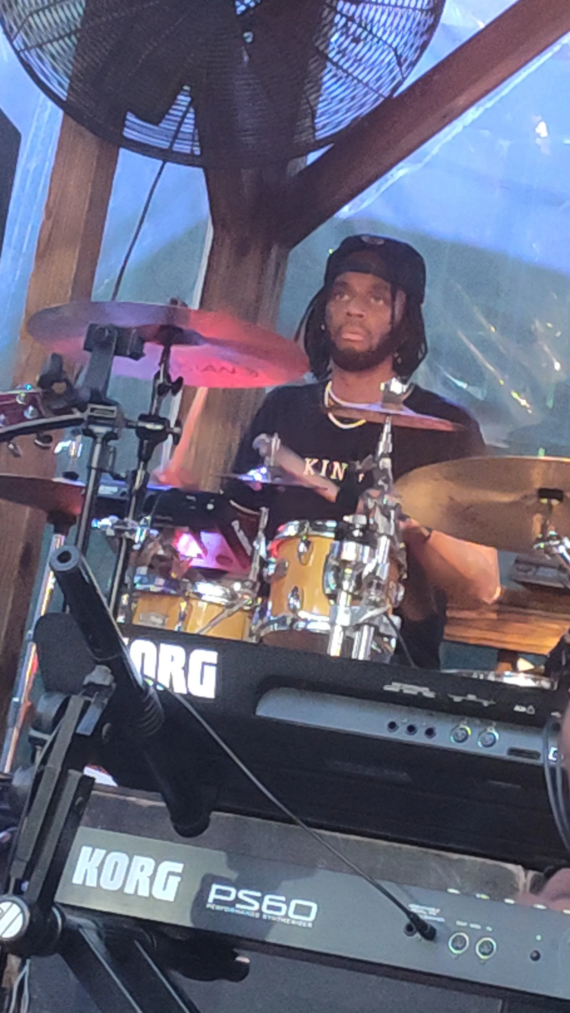 A man playing a drum set on stage, wearing a black cap and shirt, with a large fan behind him and electronic equipment in the foreground.