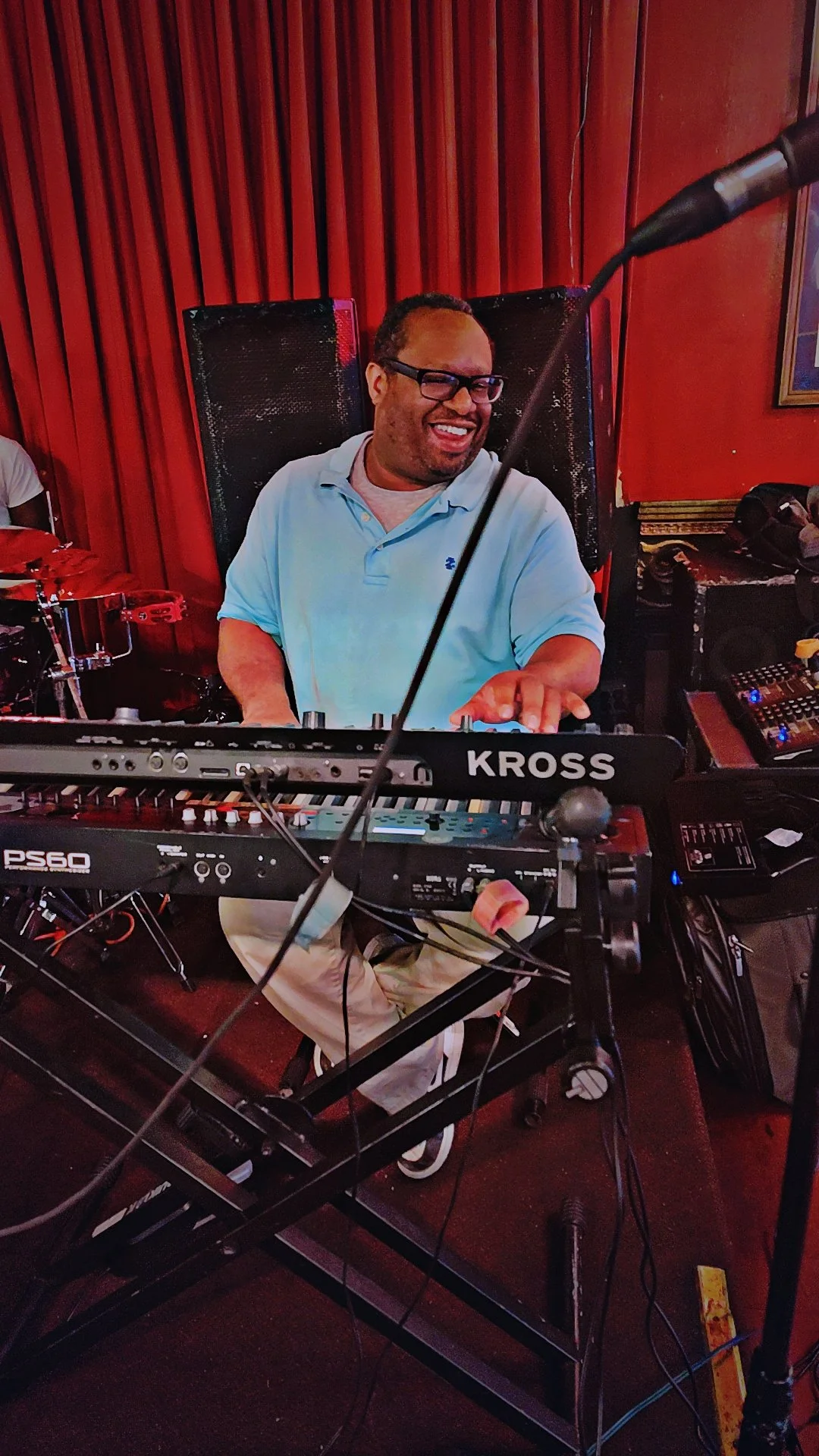 A man smiling and playing a keyboard at a music event, with red curtains in the background.