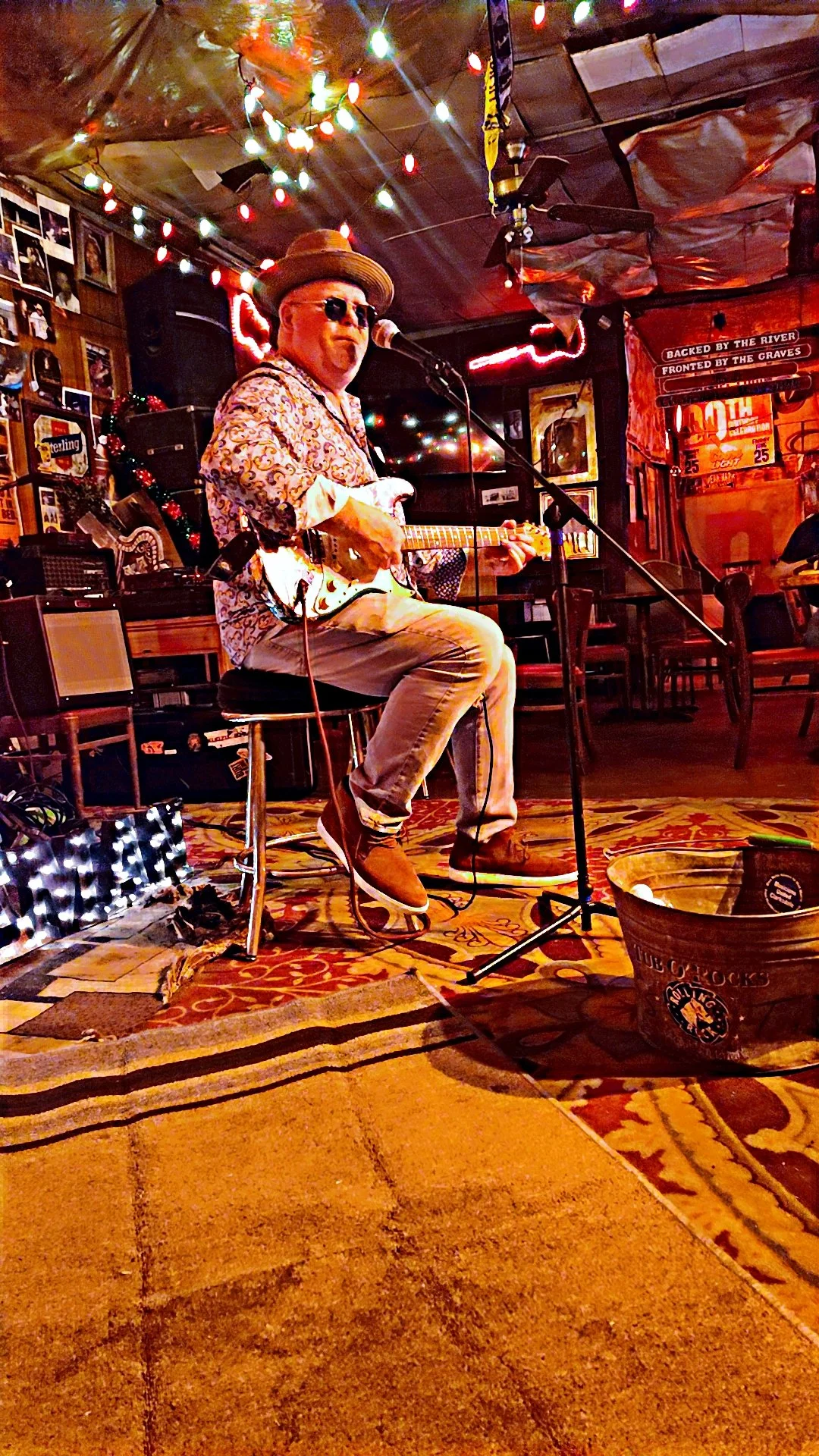 A man wearing sunglasses, a hat, and a patterned shirt playing an electric guitar on a small stage with colorful string lights in a rustic bar or pub setting.