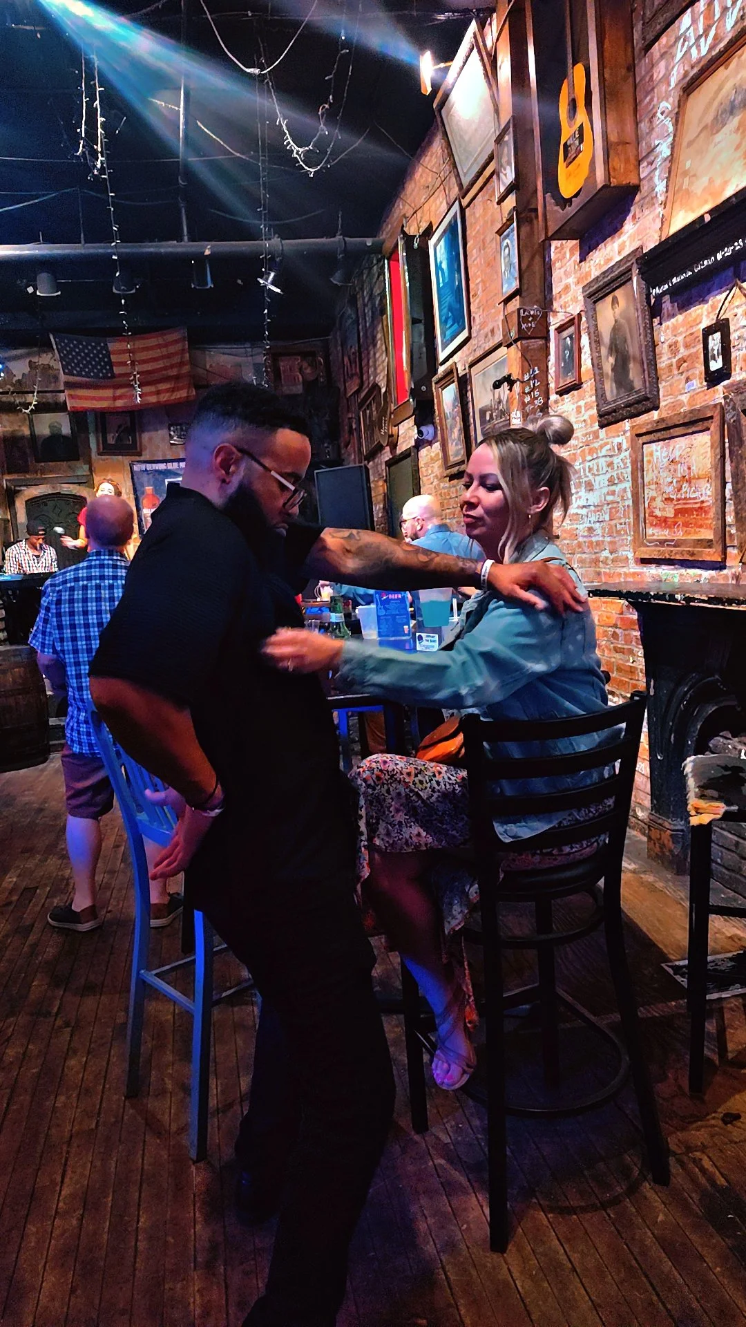 A man and a woman are dancing and sitting at a bar in a dimly lit, rustic pub decorated with framed pictures and an American flag.