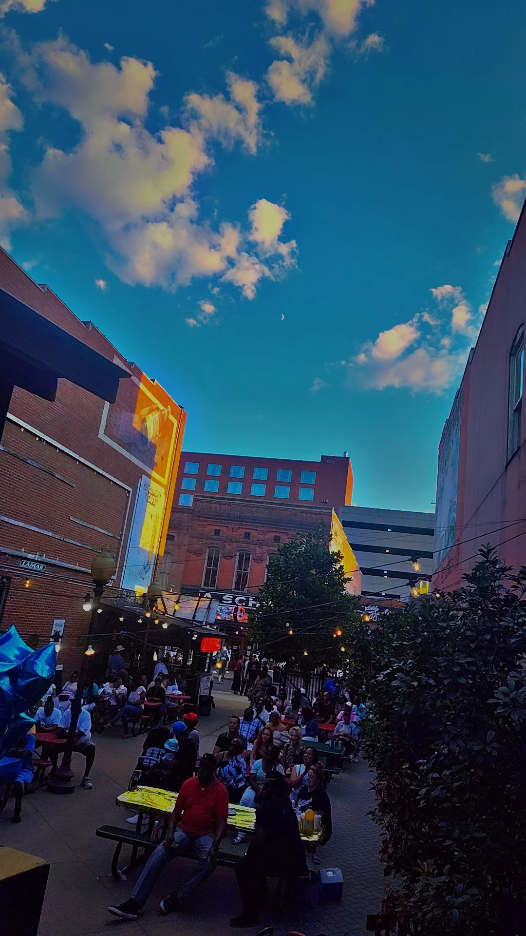 Outdoor gathering in a city plaza with people sitting at tables and balloons, surrounded by tall buildings under a partly cloudy sky with a faint moon.