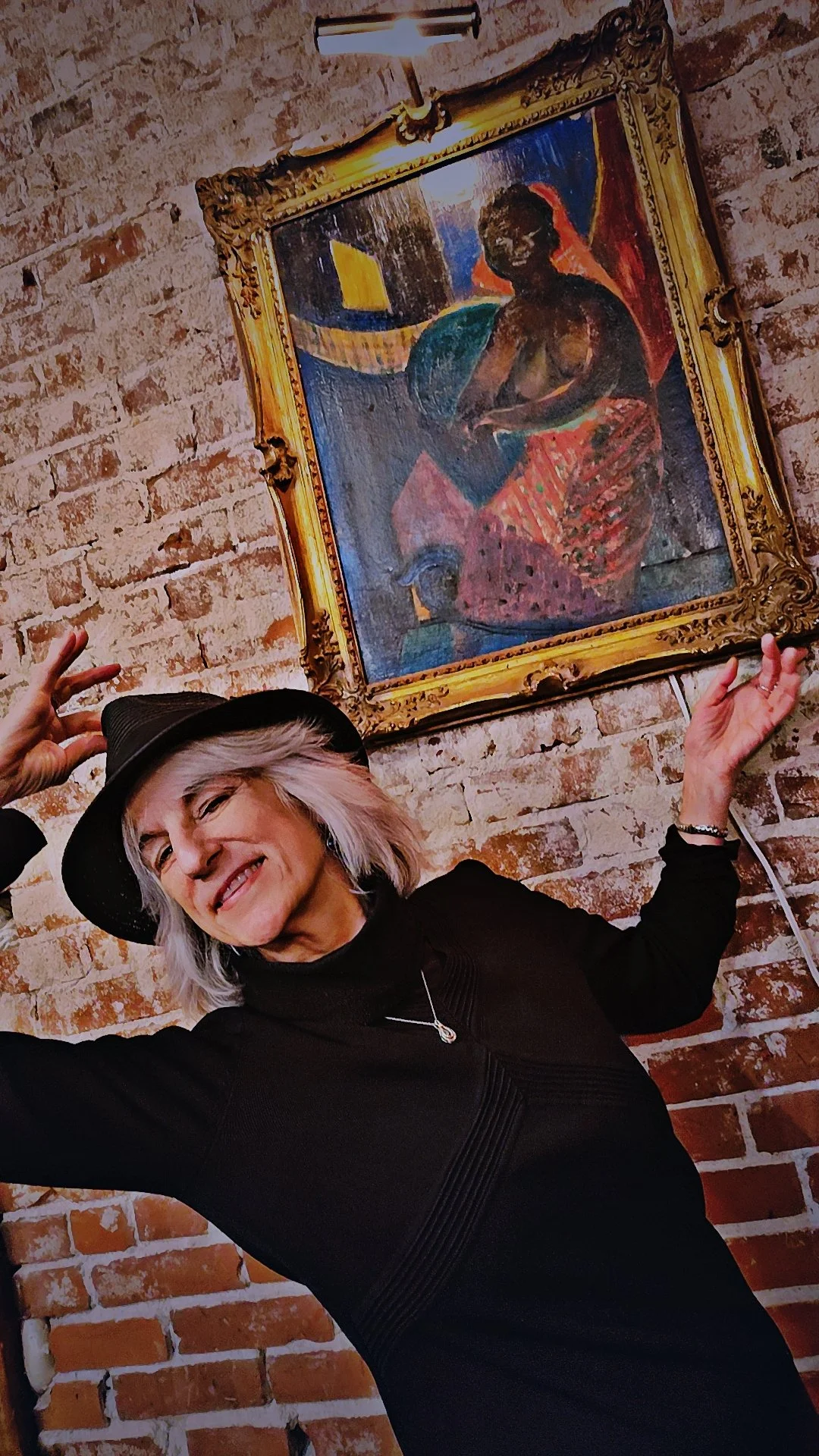 A woman with gray hair, wearing a black dress and a black hat, smiling and making a peace sign while standing next to a framed painting of a person on a brick wall.