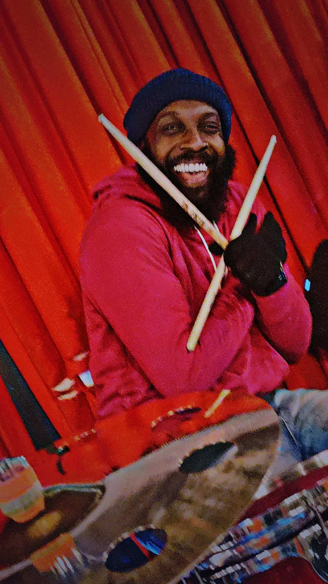 A smiling man with a beard and mustache wearing a blue beanie, a red jacket, and black gloves, holding drumsticks crossed in front of a wooden wall.