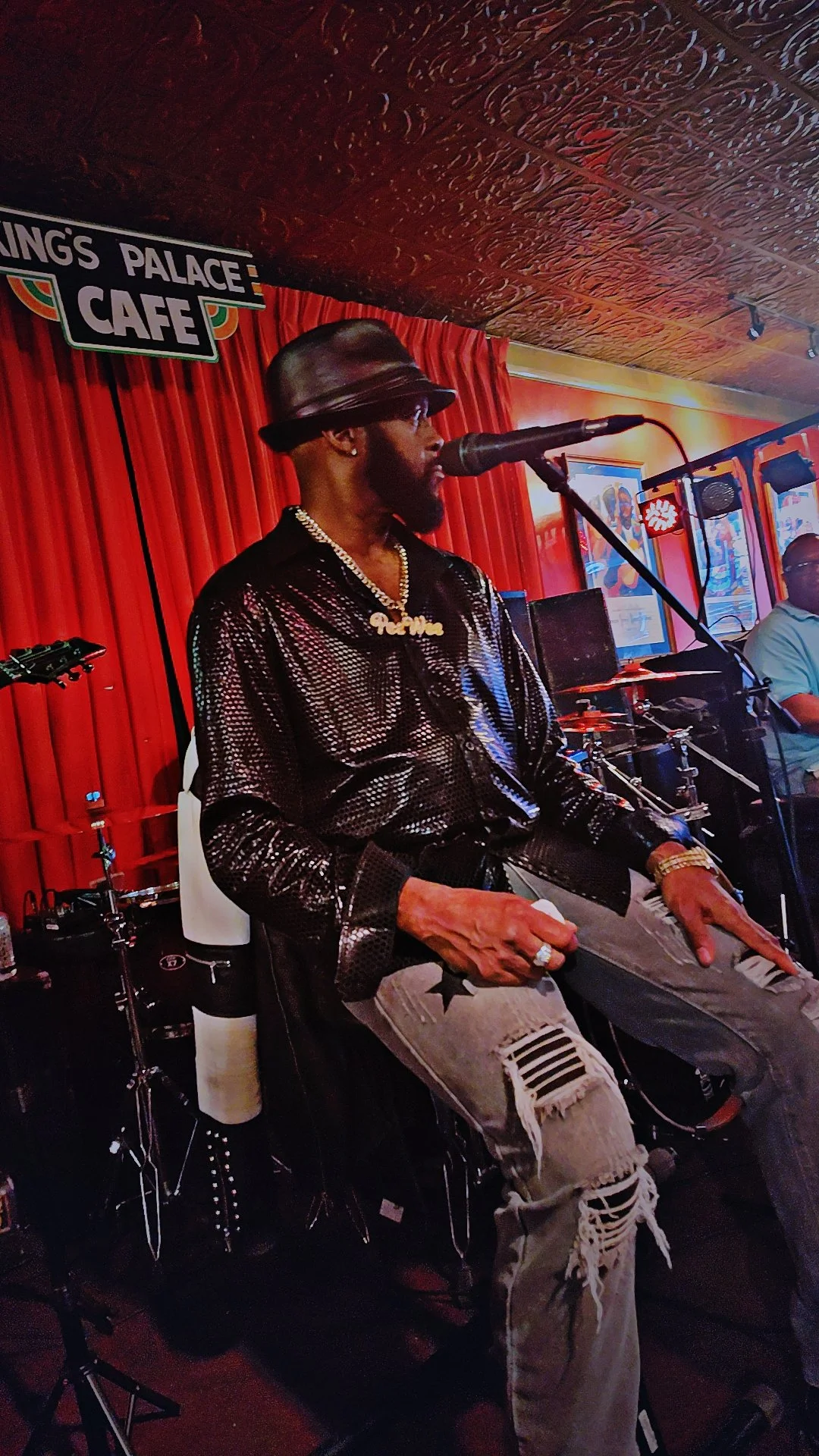 A man singing into a microphone on a stage at a lounge or cafe, wearing a black shiny jacket, ripped jeans, a hat, and jewelry, with drums and other musical equipment in the background.