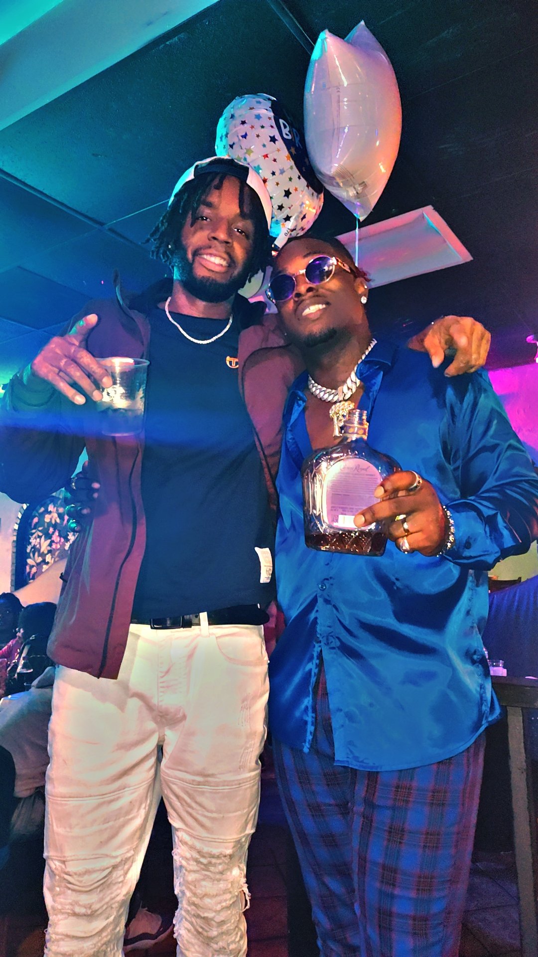Two young men celebrating in a dimly lit room with balloons in the background. One is holding a glass and wearing a dark jacket, beige distressed pants, and a white cap. The other is holding a whiskey bottle, wearing a shiny blue shirt, plaid pants, 