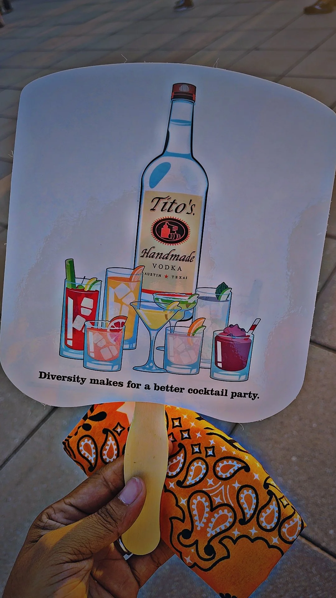 A sign promoting Tito's handmade vodka and various colorful cocktails with the slogan "Diversity makes for a better cocktail party" held on a hand with a bandana.