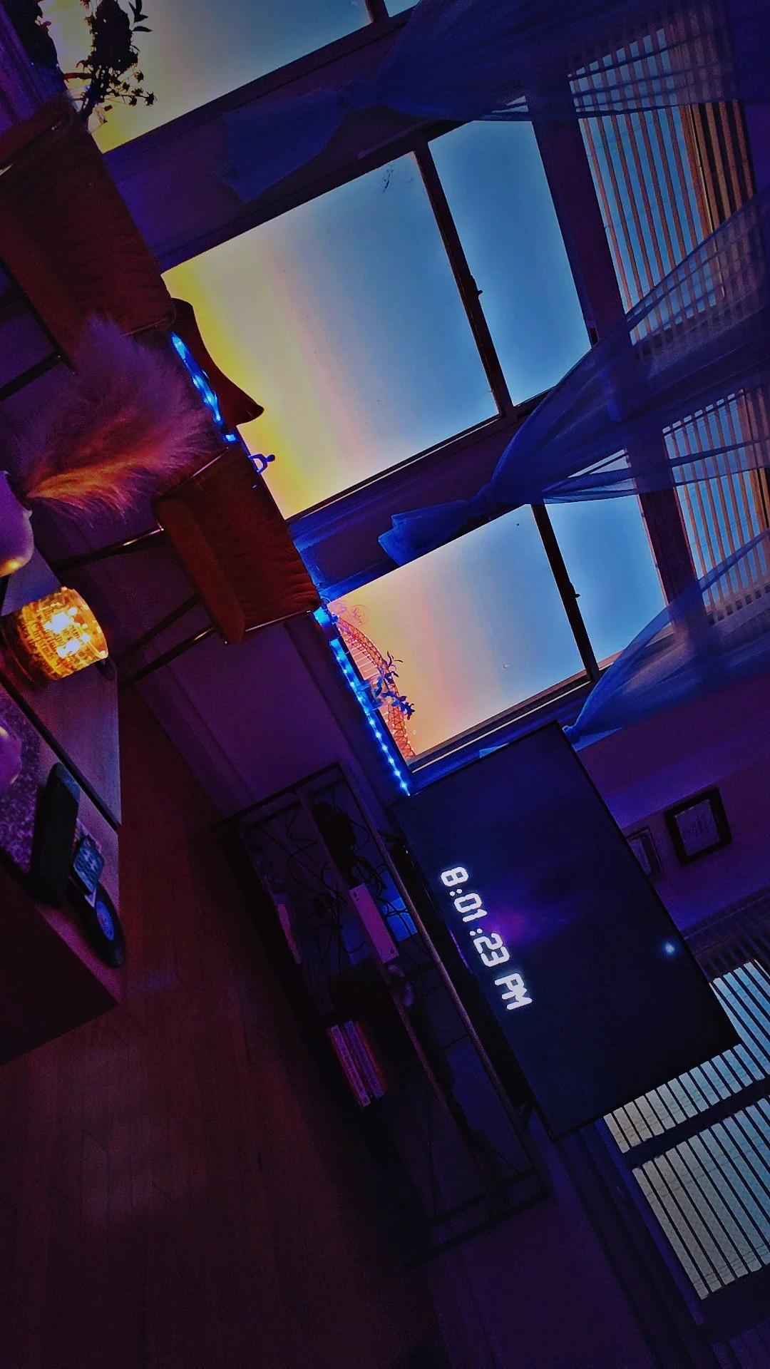 Living room with a large window showing a colorful sunset sky, decorated with blue string lights, a TV displaying the time 8:01 PM, and a wooden table with remote controls and a candle.