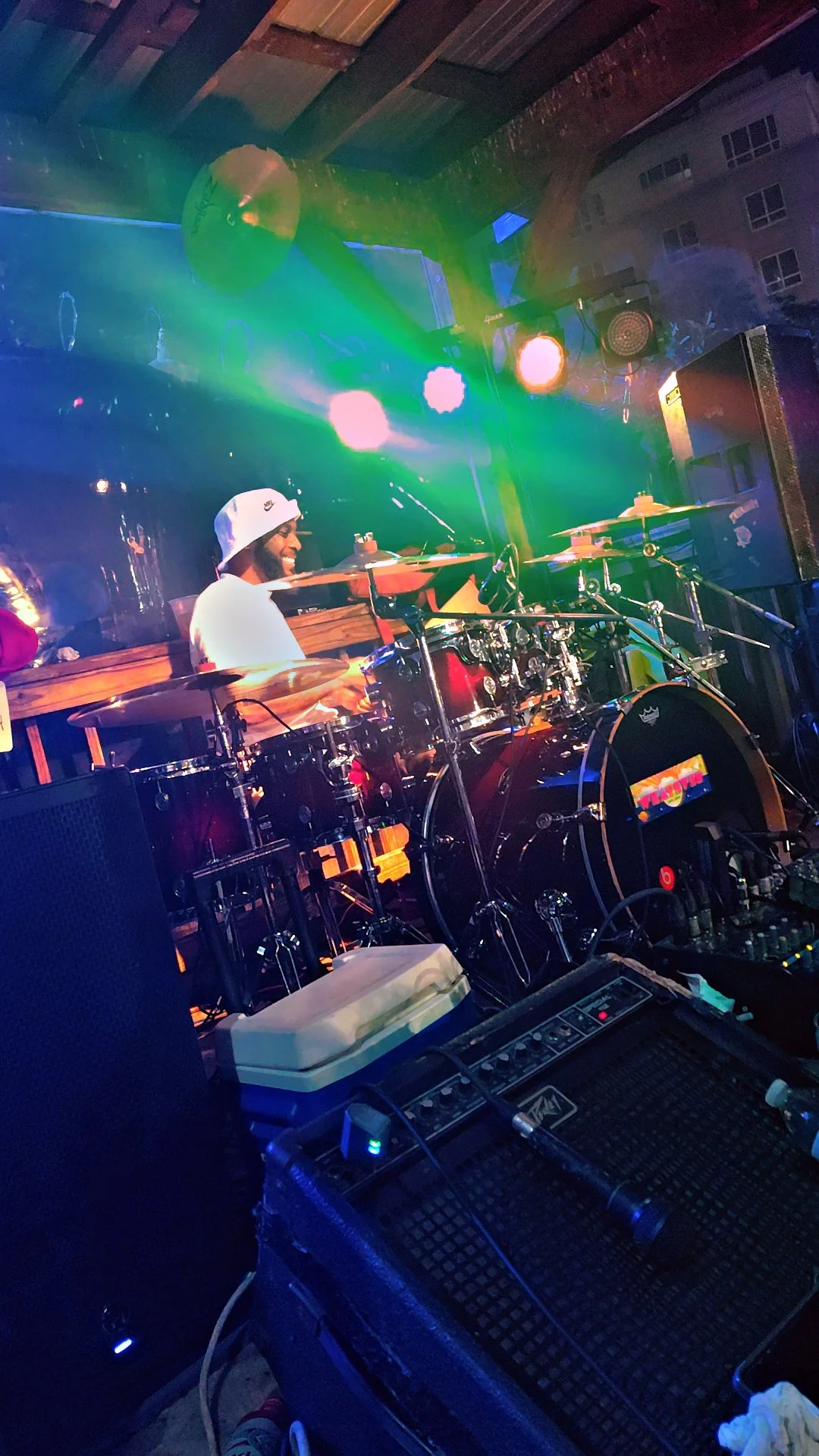 A musician playing drums on stage, smiling, with colorful stage lights illuminating the scene.