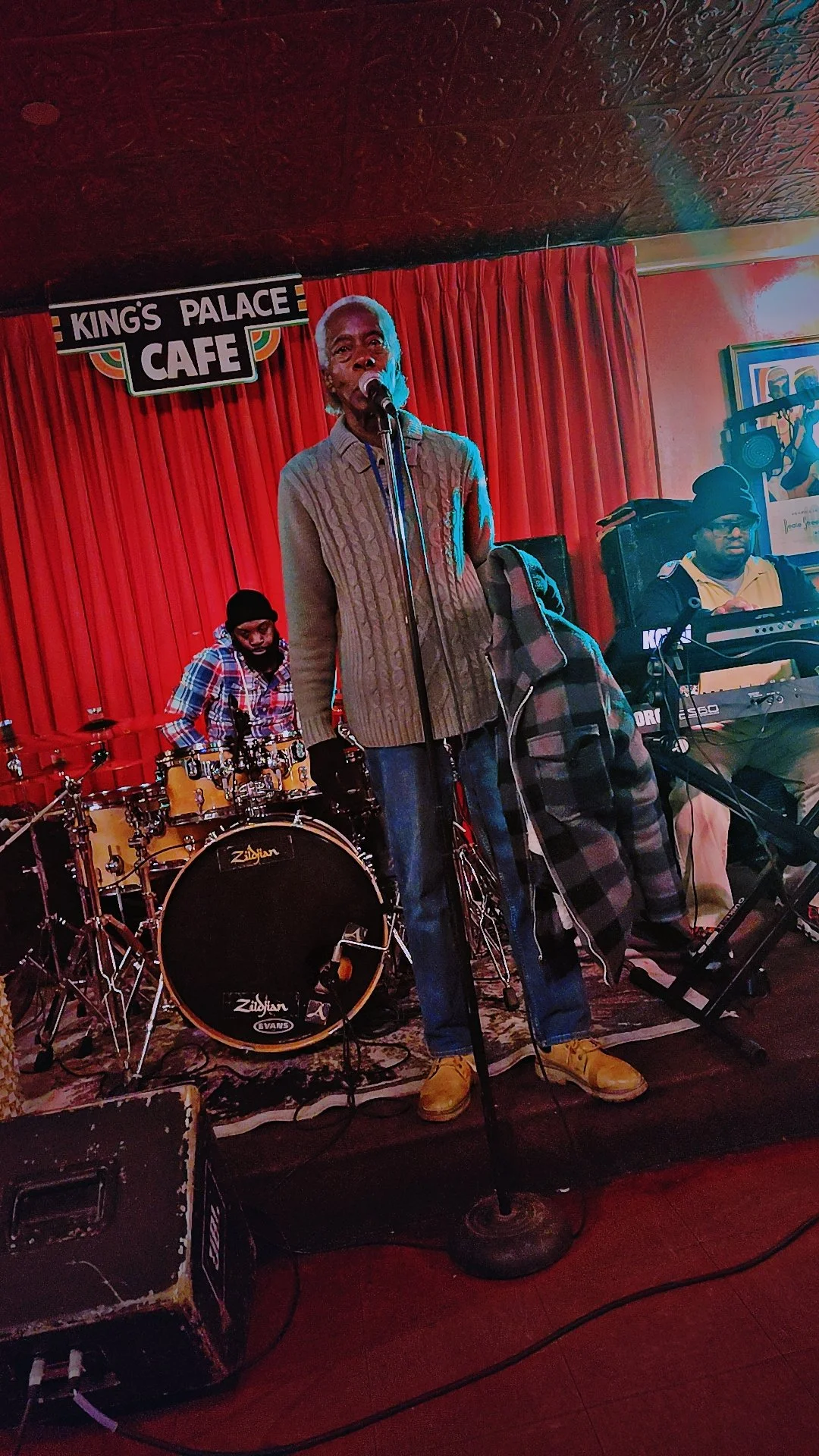 A live music performance at King's Palace Cafe featuring a singer with a microphone, a drummer, and a keyboard player on a stage with a red curtain backdrop.