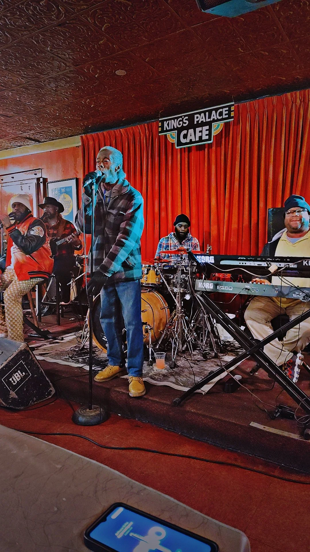 A live band performing on stage at King's Palace Cafe, with a singer in a plaid jacket and yellow shoes, a drummer, a keyboard player, and two other instrumentalists in front of red curtains.