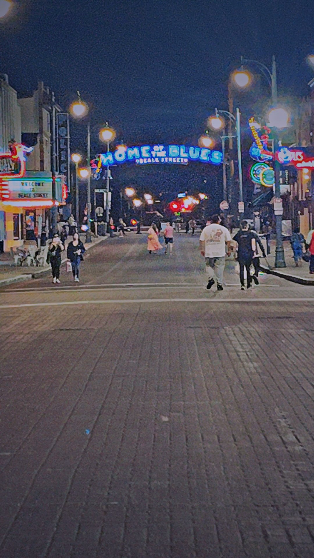 Night view of a busy street with people walking, colorful neon signs on buildings, and a sign reading "HOME OF THE BLUES" overhead.