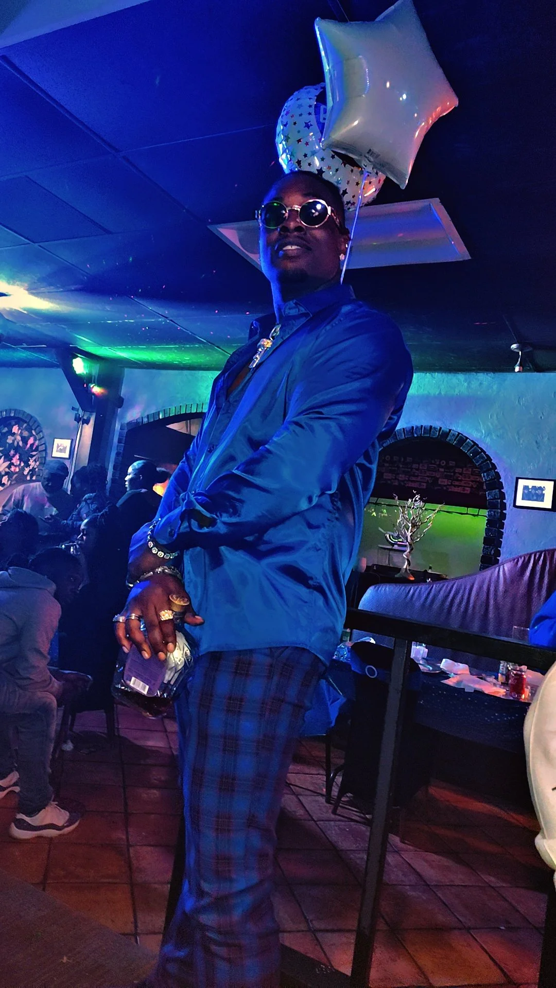 A man wearing sunglasses, a shiny blue jacket, and plaid pants standing in a dimly lit, colorful bar or club with balloons and other patrons in the background.