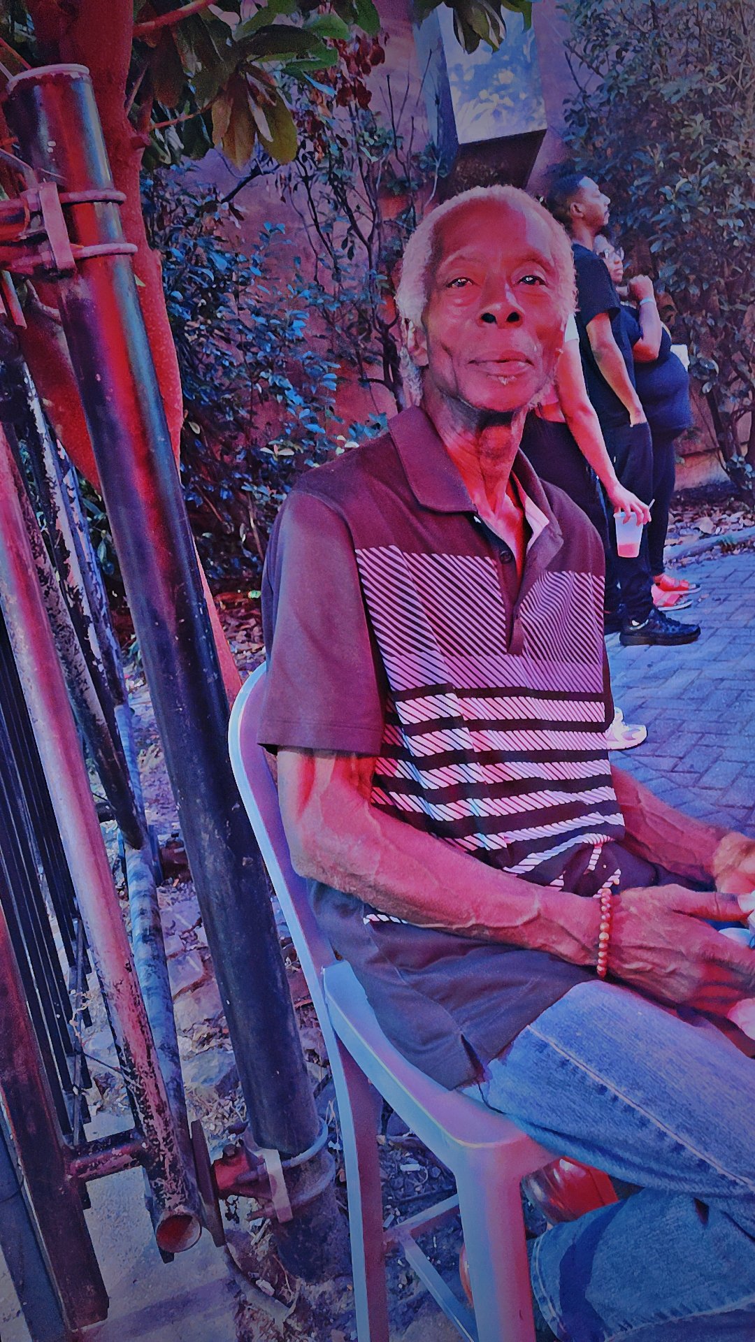 An elderly man with gray hair sitting on a white plastic chair outdoors, wearing a striped polo shirt and jeans, with bushes and other people in the background.
