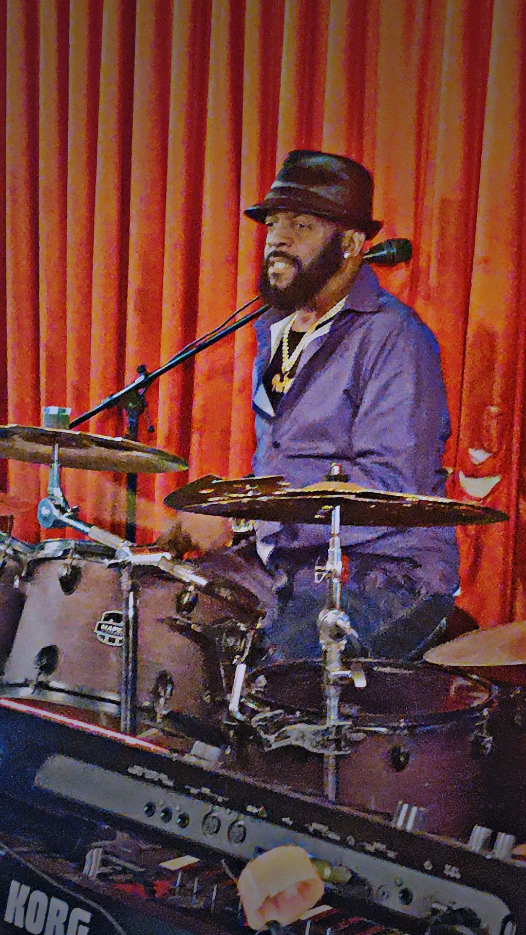 A musician playing drums and singing into a microphone on stage with red curtains in the background.