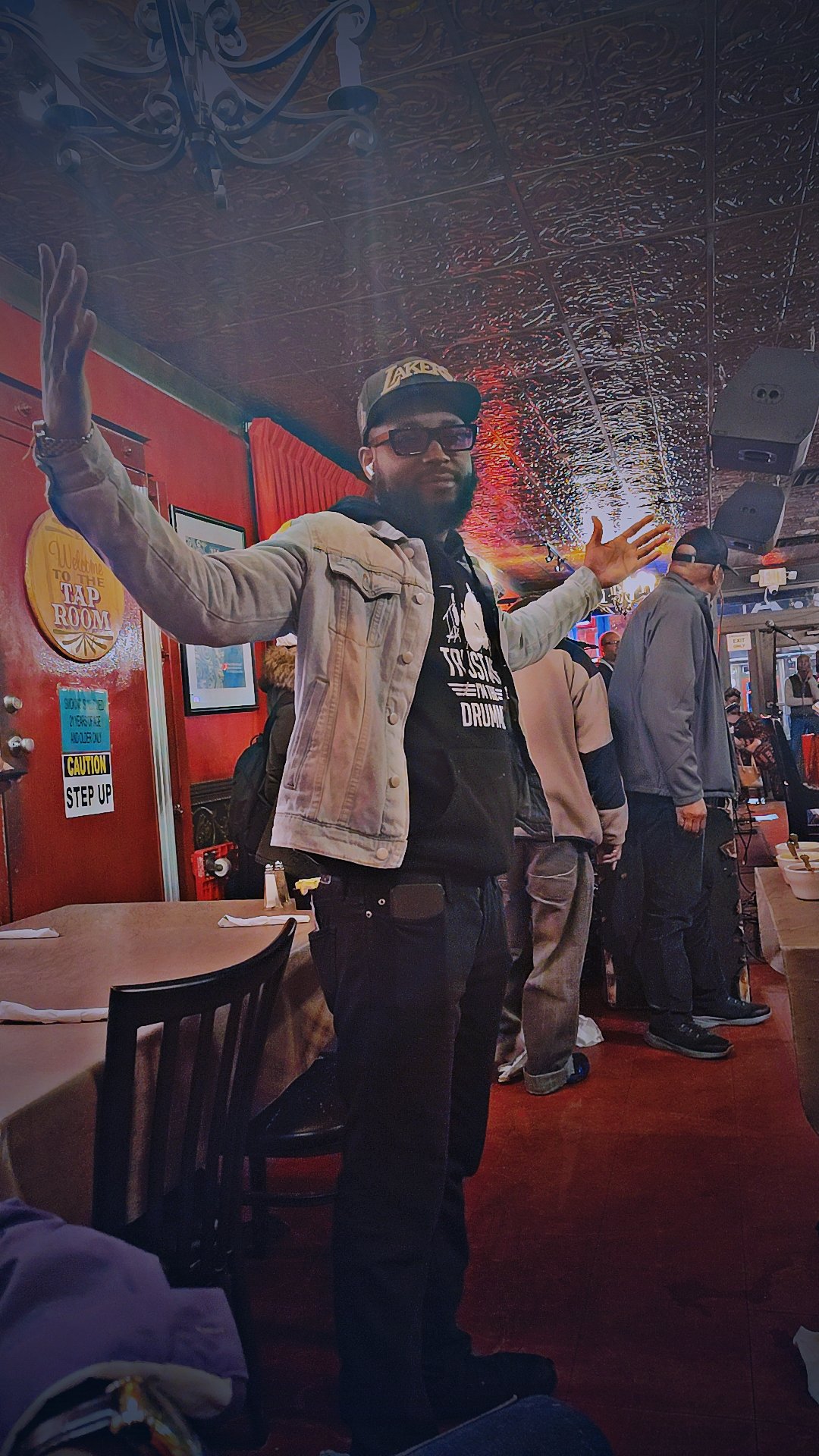 A man stands with arms outstretched inside a restaurant or bar, wearing sunglasses, a beige jacket, and a black hoodie. Several people are behind him, some with hats, and a red wall with a sign and framed pictures decorate the background.