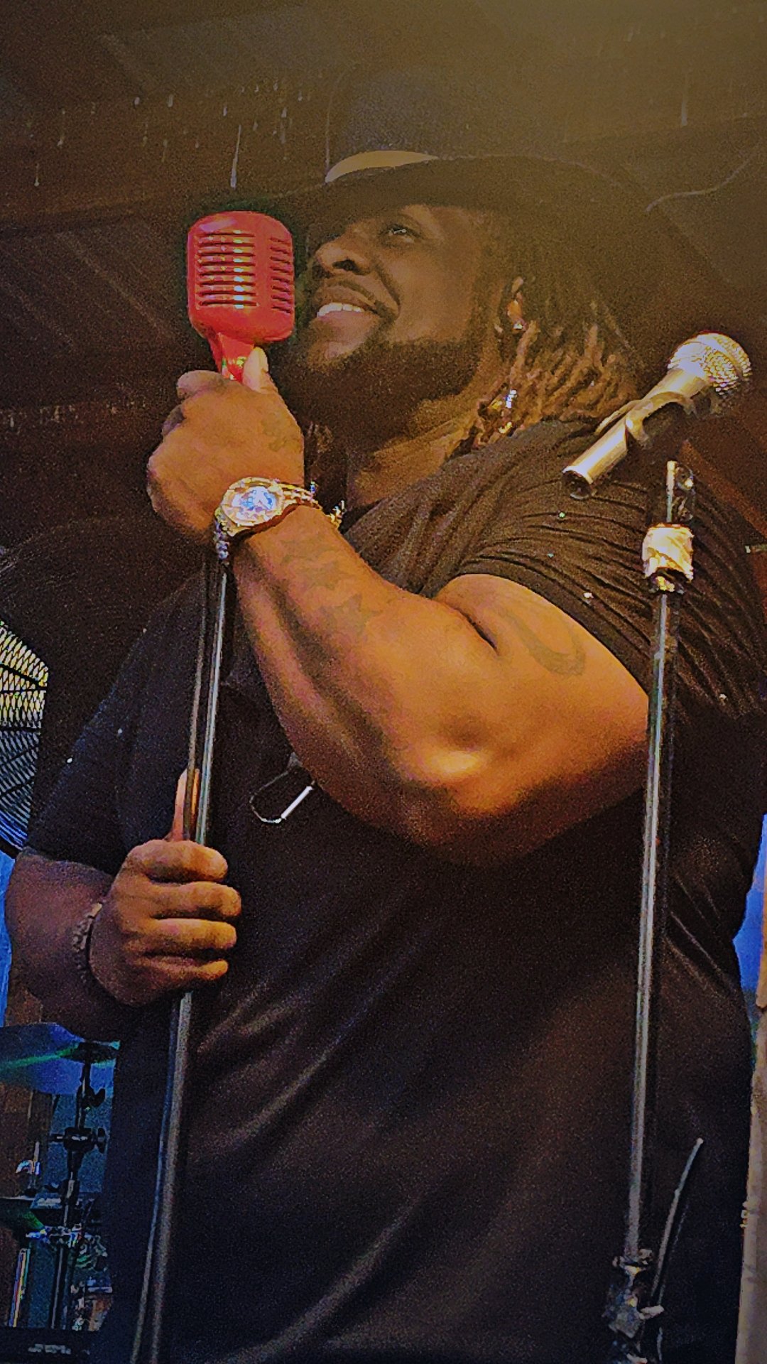 A man with a beard and dreadlocks holding a red retro microphone, smiling, with a watch on his left wrist. He is wearing a black shirt and a black wide-brimmed hat, on a stage with musical equipment visible in the background.