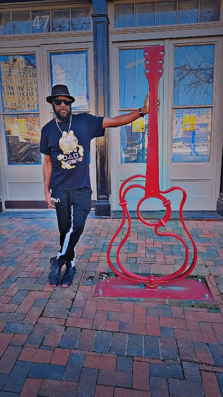 Man standing on brick sidewalk, wearing black hat, sunglasses, black T-shirt with colorful design, black pants with white stripe, black shoes, and gold chains, next to large red guitar-shaped sculpture outside storefront.