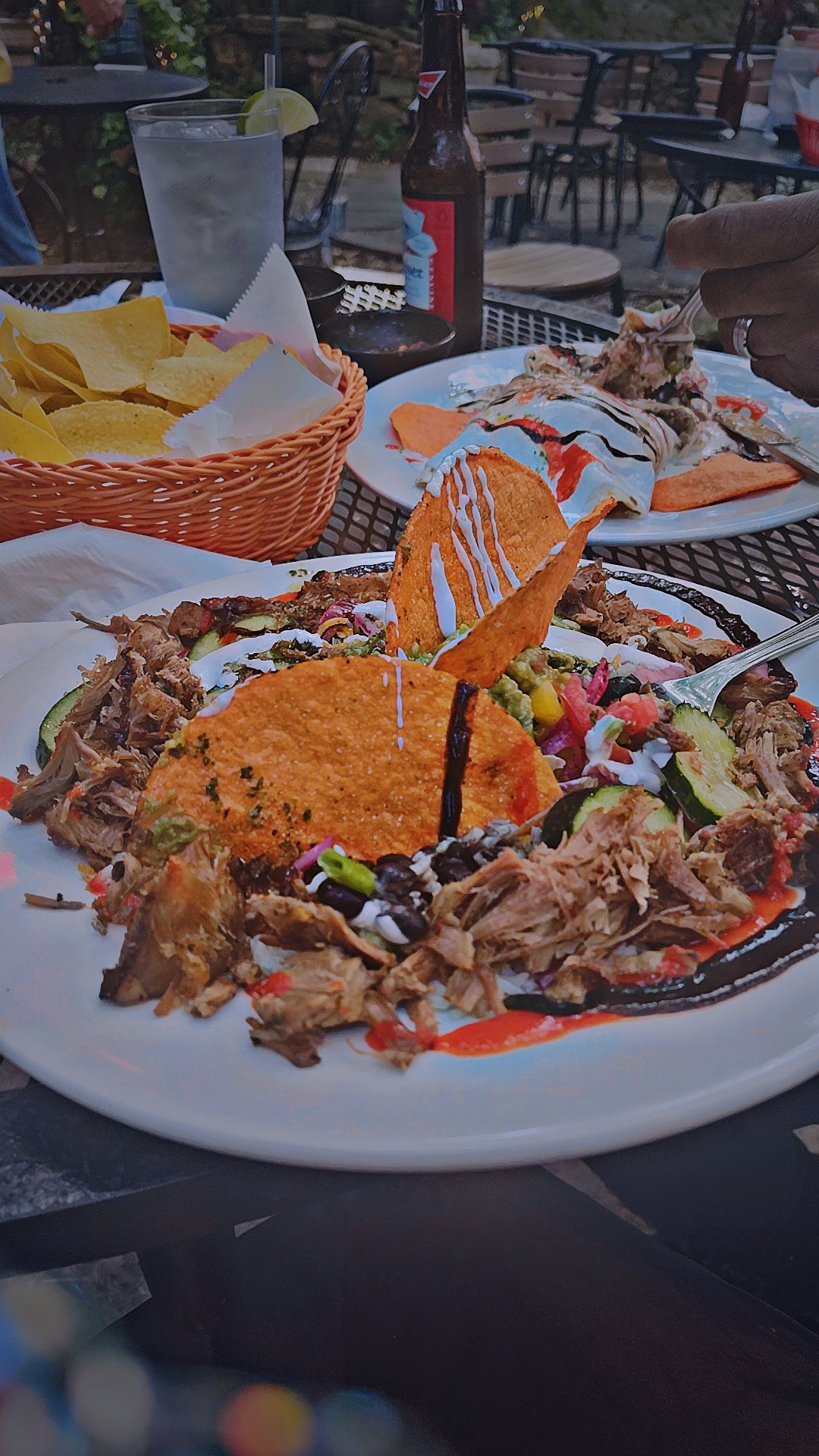 A plate of shredded meat, sliced cucumbers, diced tomatoes, and tortilla chips with drizzles of white and dark sauces, garnished with colorful vegetables, on an outdoor table.