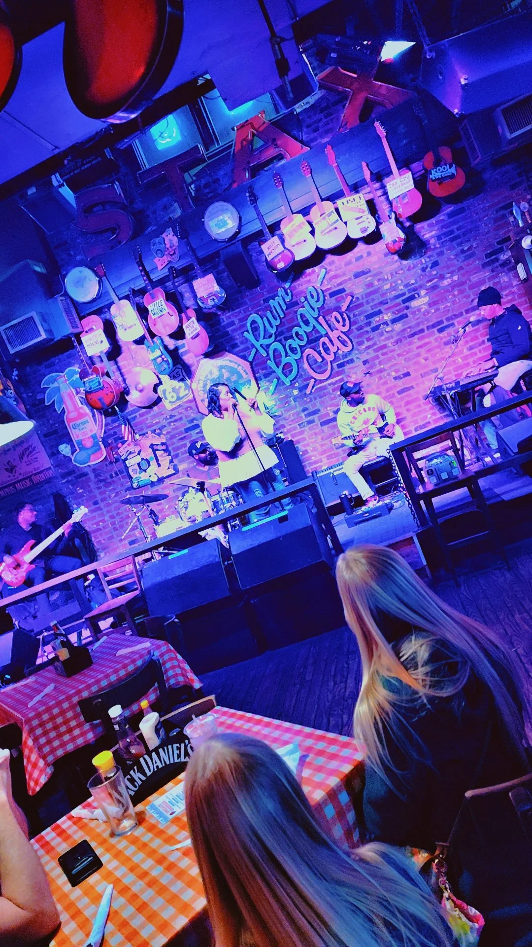 Live music performance at a bar called Rum Bar, with guitars hanging on the brick wall behind the stage and a woman singing into a microphone.
