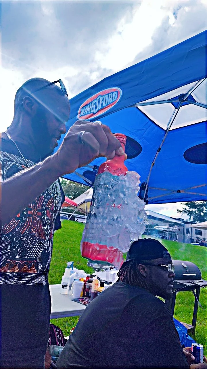 Two men at an outdoor event under a blue canopy tent, with one man holding a red drink covered in ice, and the others sitting nearby. There are various bottles and supplies on a table behind them.