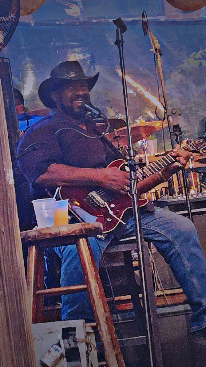 A man wearing a cowboy hat smiling while playing an electric guitar on stage with microphone stands around him, set in a rustic, lively venue with colorful lighting.